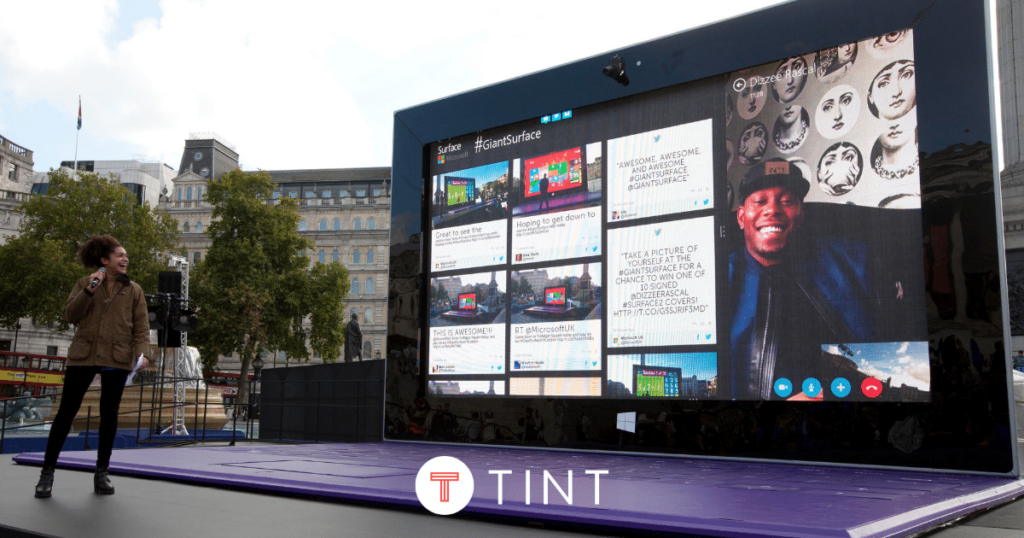 Woman with microphone standing in front of giant microsoft surface laptop, TrueLoyalmix from TrueLoyal social displays is on screen.